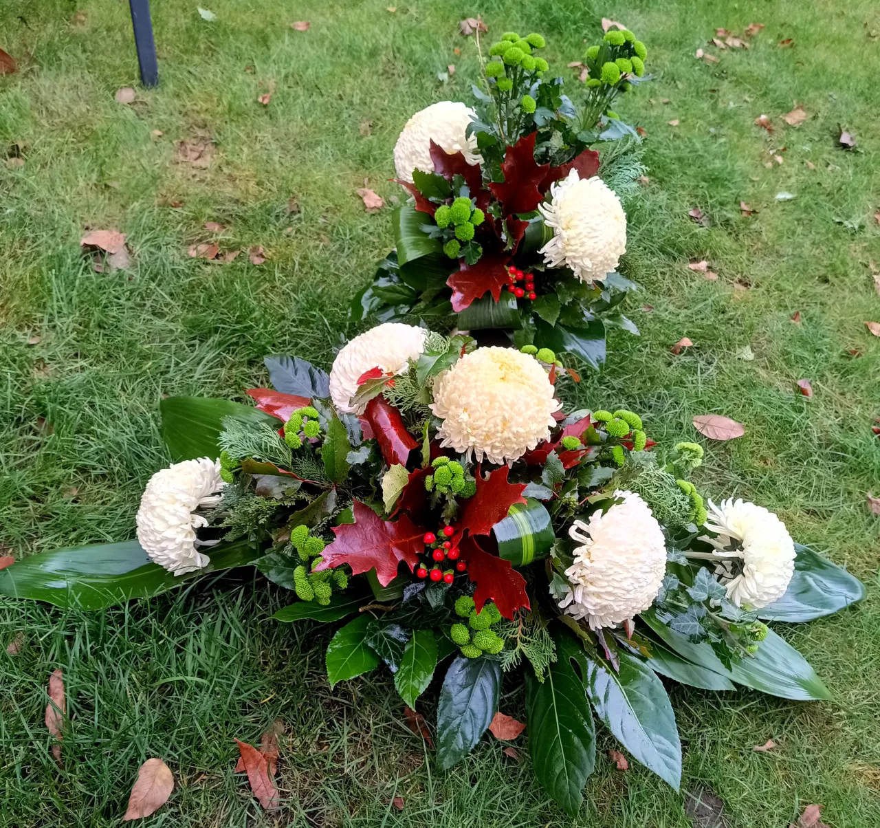 Podłużna nagrobna kompozycja z białych i zielonych chryzantem z liśćmi i czerwonym hypericum – DN 18